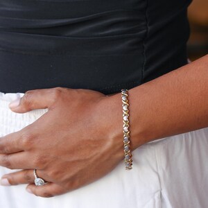 Puede incluir: Una pulsera dorada con eslabones en forma de X y O alternados, cada uno engastado con una peque&ntilde;a piedra preciosa transparente. La pulsera se lleva en la mu&ntilde;eca, con una parte superior negra y una falda blanca en el fondo. Un anillo de diamantes es visible.