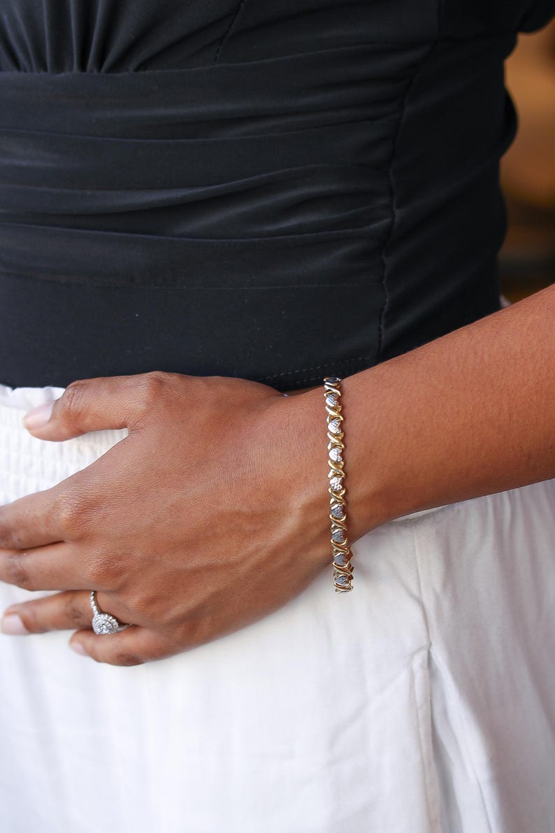 Puede incluir: Una pulsera dorada con eslabones en forma de X y O alternados, cada uno engastado con una peque&ntilde;a piedra preciosa transparente. La pulsera se lleva en la mu&ntilde;eca, con una parte superior negra y una falda blanca en el fondo. Un anillo de diamantes es visible.