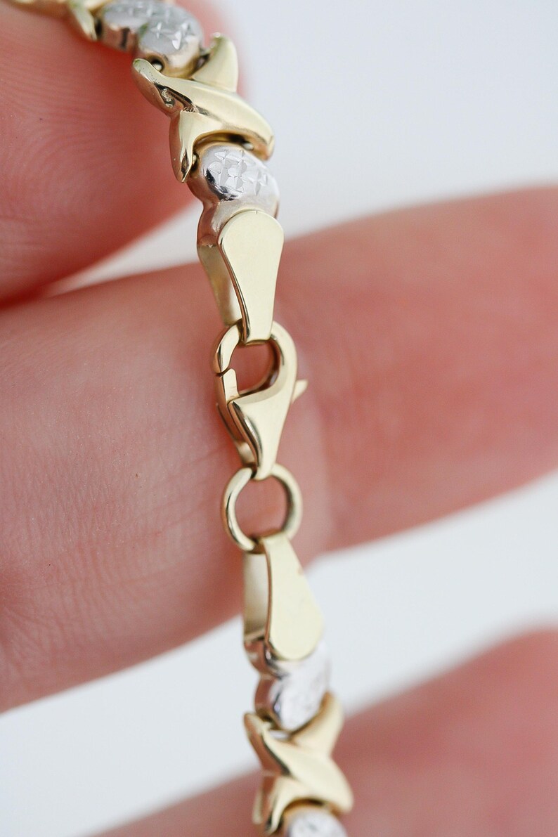 Puede incluir: Una pulsera dorada y plateada con un dise&ntilde;o de X y O. La pulsera tiene un cierre de mosquet&oacute;n. La pulsera est&aacute; hecha de metal dorado y plateado. La pulsera es una joya.