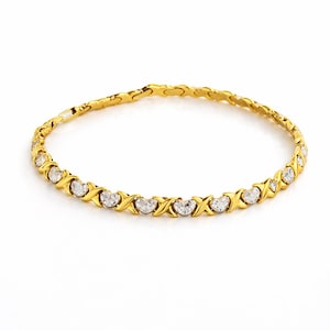 Puede incluir: Una pulsera dorada con un patr&oacute;n repetido de X y O, cada uno con una piedra transparente en forma de coraz&oacute;n. La pulsera est&aacute; sobre un fondo blanco, resaltando el dise&ntilde;o y el brillo de la joya.