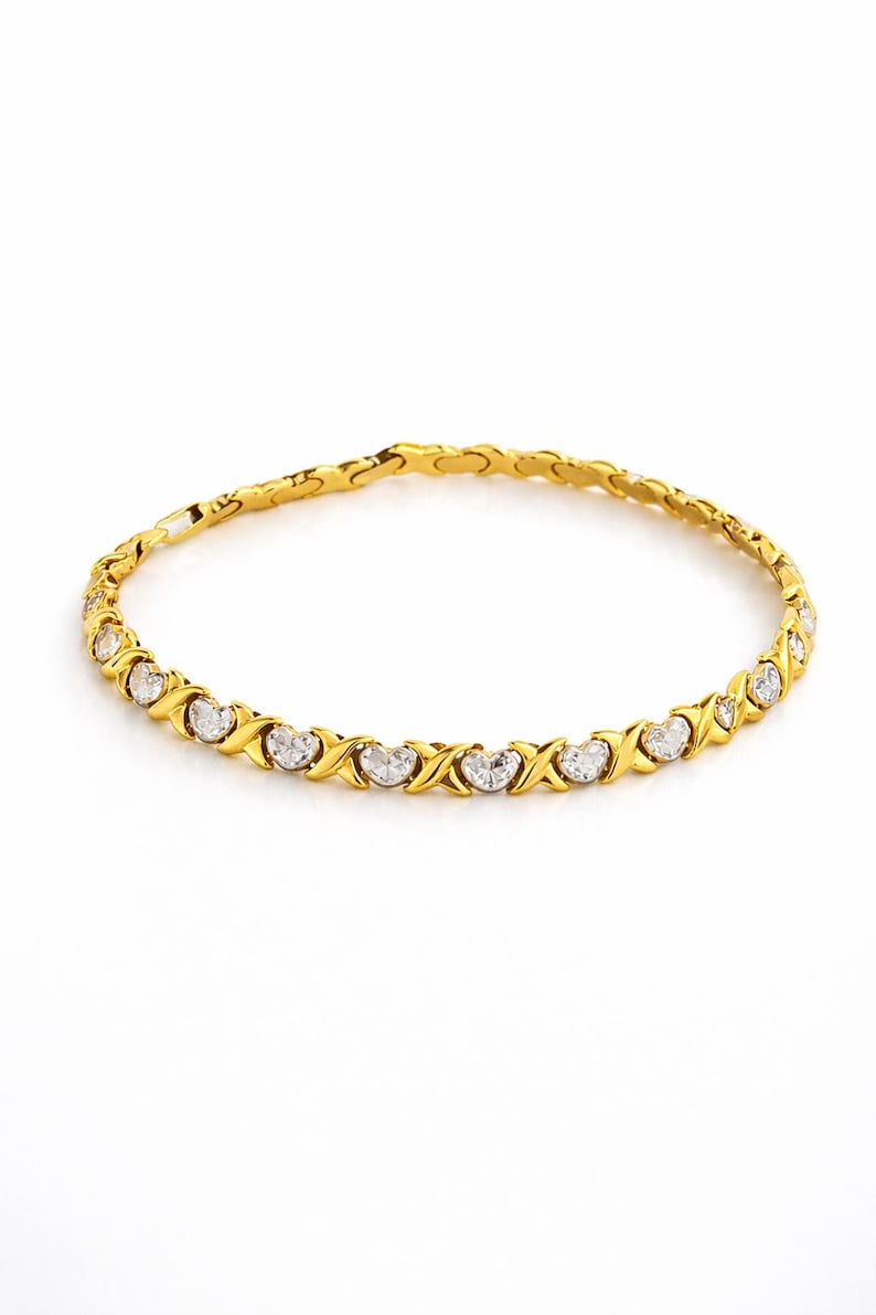 Puede incluir: Una pulsera dorada con un patr&oacute;n repetido de X y O, cada uno con una piedra transparente en forma de coraz&oacute;n. La pulsera est&aacute; sobre un fondo blanco, resaltando el dise&ntilde;o y el brillo de la joya.