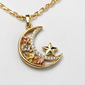 Op de afbeelding: Een gouden ketting met een halvemaanvormige hanger versierd met een rij kleine heldere steentjes en vier gestructureerde bloemaccenten in geel-, rosé- en witgoud. Een vijfpuntige gouden ster is bevestigd aan de binnenkant van de maan.