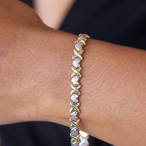 Puede incluir: Pulsera dorada con eslabones en forma de X y coraz&oacute;n alternados. El dise&ntilde;o de la pulsera incorpora una mezcla de metal dorado y plateado, creando un efecto bicolor. La pulsera se muestra en una mu&ntilde;eca, con un fondo oscuro.