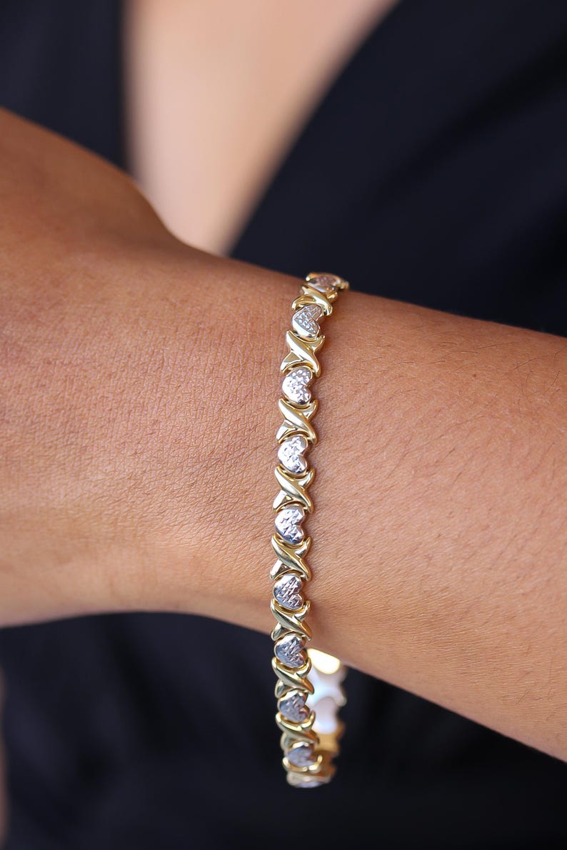 Puede incluir: Pulsera dorada con eslabones en forma de X y coraz&oacute;n alternados. El dise&ntilde;o de la pulsera incorpora una mezcla de metal dorado y plateado, creando un efecto bicolor. La pulsera se muestra en una mu&ntilde;eca, con un fondo oscuro.