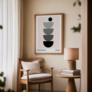 Könnte beinhalten: Ein gerahmter Druck mit einem geometrischen Design aus übereinander gestapelten Halbkreisen in Grautönen und Schwarz auf beigefarbenem Hintergrund. Der Druck trägt den Text "BAUHAUS 1923" unten. Der Druck hängt an einer Wand neben einem Holzstuhl und einer Lampe.