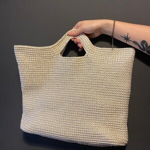 May include: A beige crocheted tote bag with a woven texture. The bag has a single handle with a cut-out design.