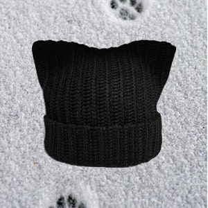 Op de afbeelding: Zwarte gebreide muts met kattenoren op een besneeuwde achtergrond met drie potafdrukken.