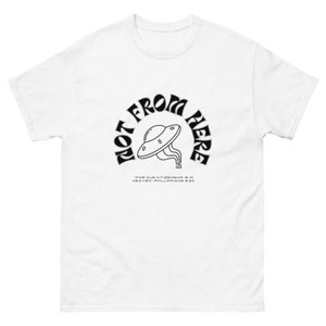 May include: White t-shirt featuring a black graphic of a UFO and the text "NOT FROM HERE" in a curved arch. Underneath the UFO, the text reads "FOR OUR CITIZENSHIP IS IN HEAVEN PHILIPPIANS 3:20".