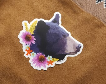 May include: A black bear head sticker with a floral wreath around it. The flowers are purple, orange, and yellow. The sticker has the text "hakes.art" on the bottom.