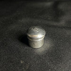 May include: A small, cylindrical silver bottle stopper with a polished, rounded top. The top surface features an intricate, engraved monogram consisting of two intertwined cursive letters. A small hallmark is visible on the side of the base.