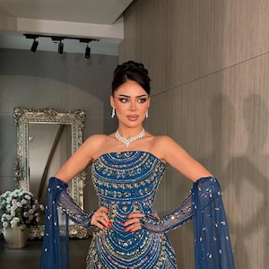 May include: A floor-length teal strapless gown featuring intricate beadwork and sequin embellishments in a fan-like pattern. The dress includes matching sheer, long, detached sleeves adorned with scattered crystals and a silver-toned necklace.