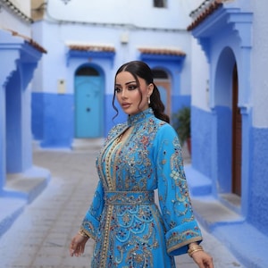 May include: A woman in a long, ornate, turquoise dress with intricate gold embroidery and jewel accents. The dress features a high collar and long sleeves. The background is a blue-walled alleyway.