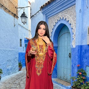 May include: A floor-length, deep red kaftan featuring intricate gold embroidery along the neckline, chest, and sleeve cuffs. The garment is paired with a layered gold statement necklace and matching gold bracelets, creating a cohesive look.