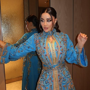 May include: A long, light blue dress with intricate gold embroidery and embellishments. The dress features a high collar, long sleeves, and a fitted waist. The design includes floral and geometric patterns, with a matching belt. The dress is reflected in a mirror.