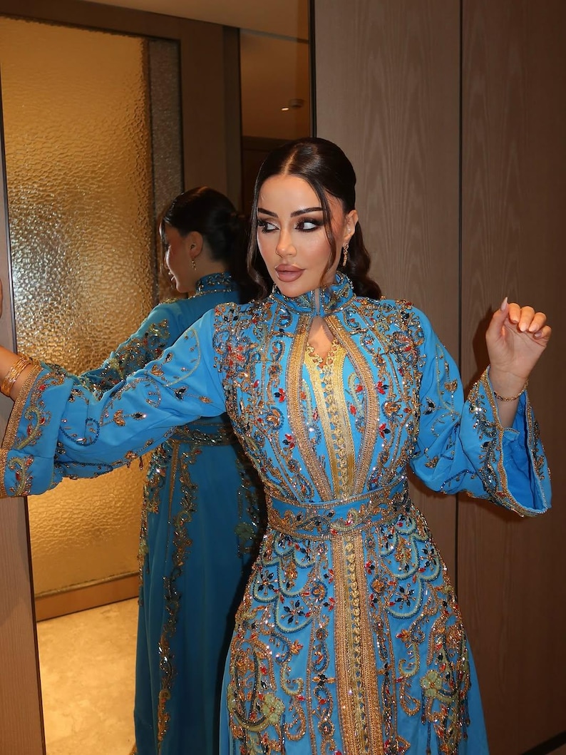 May include: A long, light blue dress with intricate gold embroidery and embellishments. The dress features a high collar, long sleeves, and a fitted waist. The design includes floral and geometric patterns, with a matching belt. The dress is reflected in a mirror.