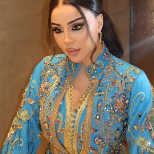 May include: A woman wearing a turquoise dress with intricate gold embroidery and embellishments. The dress features a high collar and long sleeves, with detailed patterns and sparkling accents. The overall design is ornate and elegant.