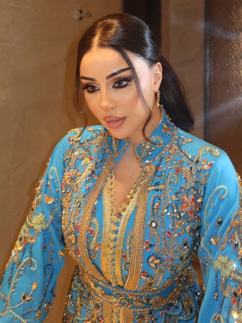 May include: A woman wearing a turquoise dress with intricate gold embroidery and embellishments. The dress features a high collar and long sleeves, with detailed patterns and sparkling accents. The overall design is ornate and elegant.
