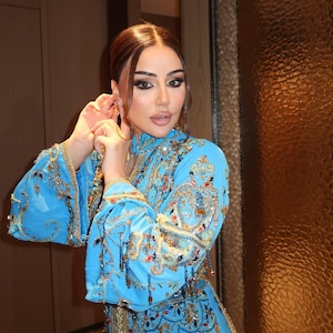 May include: A light blue, ornate dress with gold embroidery and embellishments. The dress features long sleeves and intricate patterns. The person is wearing gold earrings. The dress is a traditional garment.
