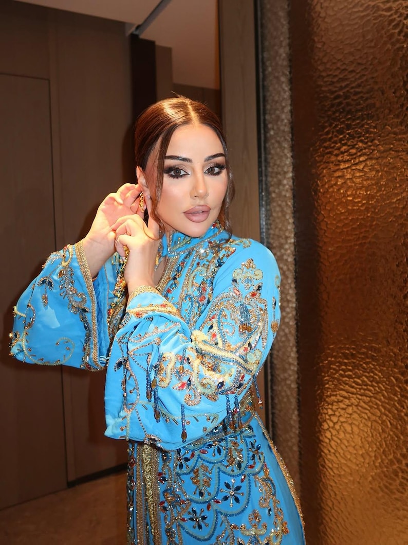May include: A light blue, ornate dress with gold embroidery and embellishments. The dress features long sleeves and intricate patterns. The person is wearing gold earrings. The dress is a traditional garment.