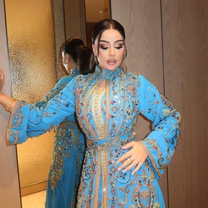 May include: A long, light blue dress with intricate gold and colorful embroidery. The dress features a high collar, long sleeves, and a fitted waist. The embroidery includes floral and geometric patterns, with sparkling embellishments.