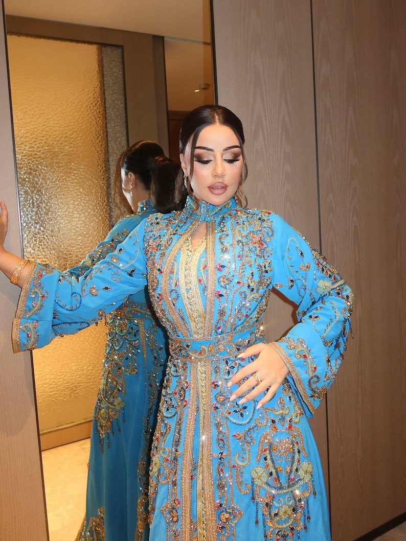 May include: A long, light blue dress with intricate gold and colorful embroidery. The dress features a high collar, long sleeves, and a fitted waist. The embroidery includes floral and geometric patterns, with sparkling embellishments.
