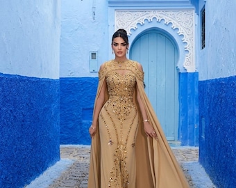 May include: A floor-length gold gown featuring cold-shoulder cutouts, a high neckline, and an attached cape. The bodice and skirt are heavily embellished with intricate gold beadwork, sequins, and metallic embroidery in a vertical pattern.