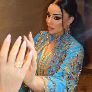 May include: A light blue embroidered caftan with gold and jewel-toned accents. The caftan features intricate patterns and embellishments. A gold ring is visible on the hand. The caftan's sleeves are long and flowing.