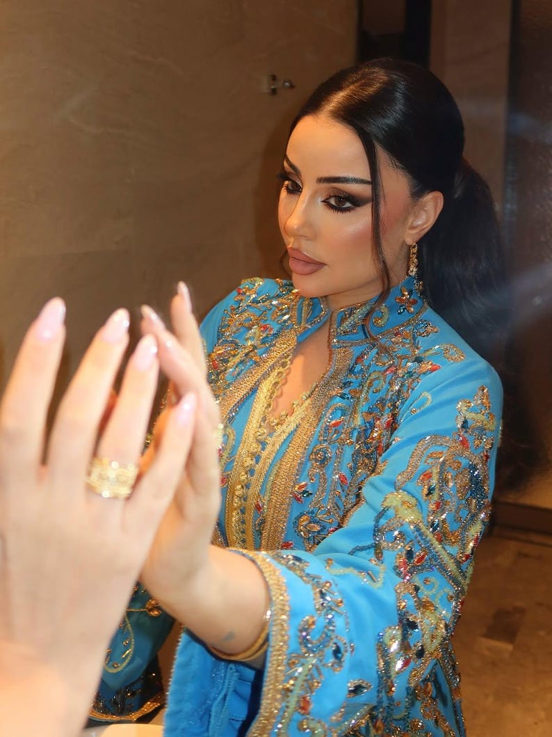 May include: A light blue embroidered caftan with gold and jewel-toned accents. The caftan features intricate patterns and embellishments. A gold ring is visible on the hand. The caftan's sleeves are long and flowing.
