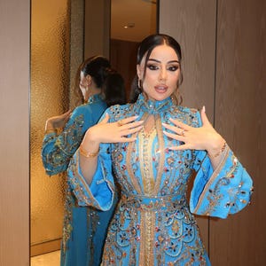May include: A turquoise blue, long-sleeved gown with intricate gold and jewel embellishments. The dress features a high neckline, flared sleeves, and a fitted waist. The gown is reflected in a mirror, showcasing the detailed design.