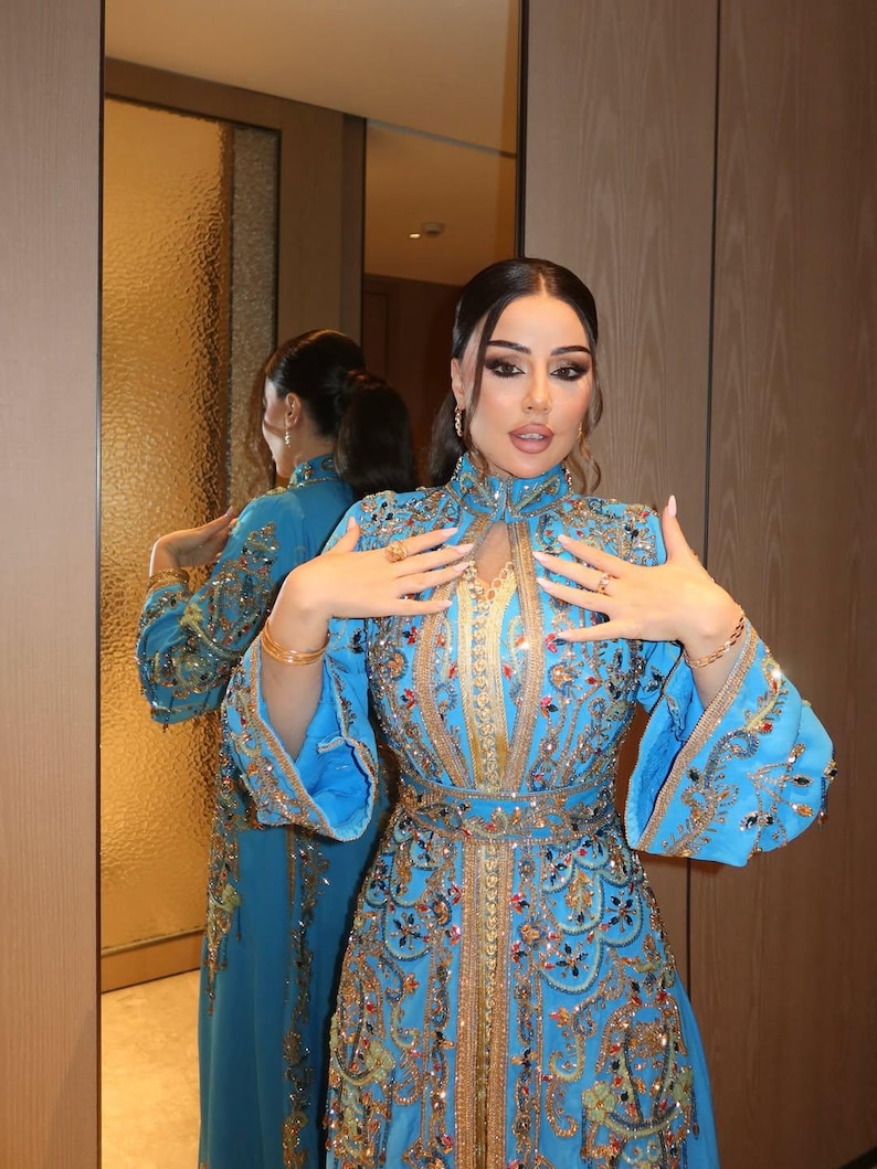 May include: A turquoise blue, long-sleeved gown with intricate gold and jewel embellishments. The dress features a high neckline, flared sleeves, and a fitted waist. The gown is reflected in a mirror, showcasing the detailed design.