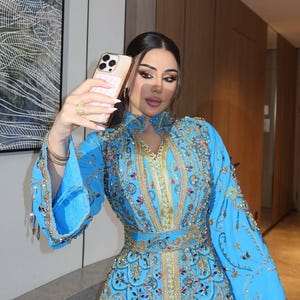 May include: A light blue, long-sleeved dress with intricate gold embroidery and embellishments. The dress features a high collar and a cinched waist. The sleeves are flared, and the dress is adorned with sequins and beads. The person is holding a gold phone.