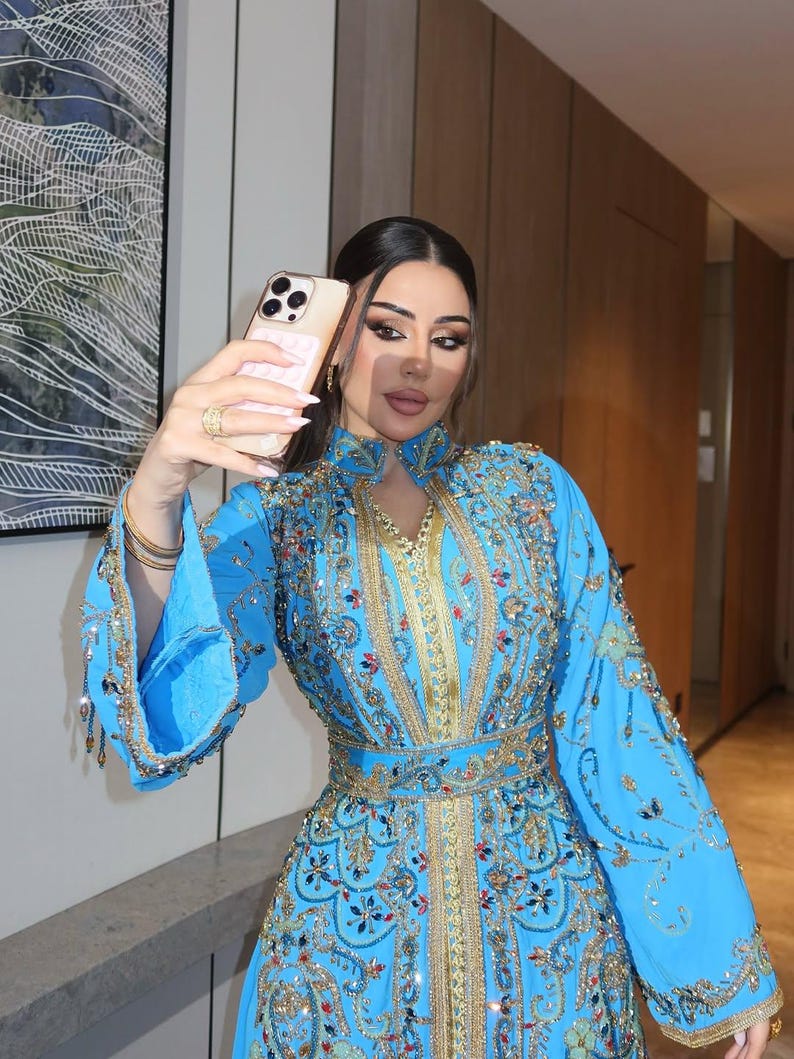 May include: A light blue, long-sleeved dress with intricate gold embroidery and embellishments. The dress features a high collar and a cinched waist. The sleeves are flared, and the dress is adorned with sequins and beads. The person is holding a gold phone.