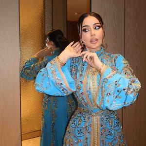 May include: A vibrant turquoise dress with intricate gold embroidery and embellishments. The dress features long sleeves, a high neckline, and a fitted bodice, reflecting a luxurious and ornate design. The dress is reflected in a mirror.