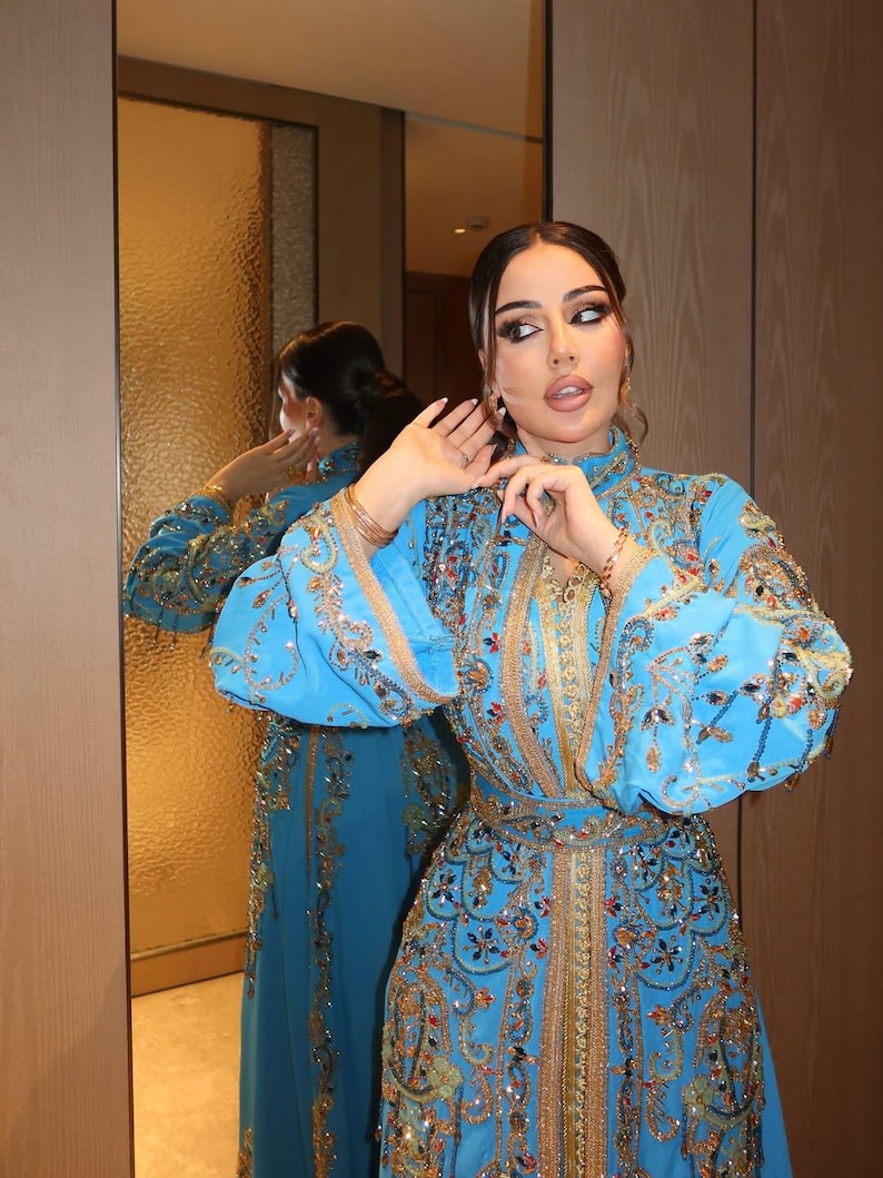May include: A vibrant turquoise dress with intricate gold embroidery and embellishments. The dress features long sleeves, a high neckline, and a fitted bodice, reflecting a luxurious and ornate design. The dress is reflected in a mirror.