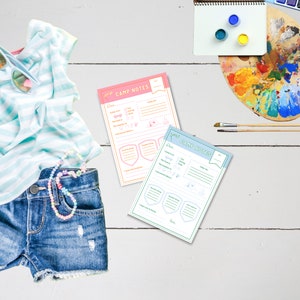 May include: Two printable camp note sheets, one pink and one blue, with a checklist of activities and space for writing. The sheets are on a white wooden surface with a pair of blue jean shorts and a blue and white striped shirt.
