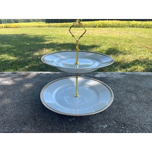 May include: Two-tiered serving stand with white plates and gold trim. The plates are supported by a gold-colored metal stand with a decorative handle. The stand is set against a backdrop of green grass and trees.