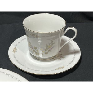 May include: White teacup and saucer set with a floral design in soft pastel colors. The cup and saucer have gold trim and a delicate pattern of flowers and leaves. The set is likely made of porcelain or fine china.