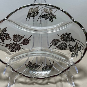 Puede incluir: Plato de servir de vidrio transparente con tres secciones y un borde plateado. El plato está decorado con diseños de hojas plateadas. El plato está sostenido por un soporte de plástico transparente.