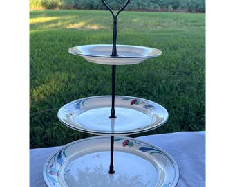 May include: A three-tiered serving stand with white plates featuring a floral and leaf design. The plates are supported by a black metal stand with a decorative handle. The stand is perfect for displaying desserts or appetizers.