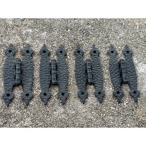 May include: Six black metal hinges with a hammered finish and decorative pointed ends. The hinges are laid out in a row on a grey gravel surface.