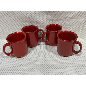 May include: Four red ceramic mugs with rounded bodies and large handles are arranged on a patterned white surface. The mugs have a glossy finish and a simple, classic design, suitable for coffee or tea.