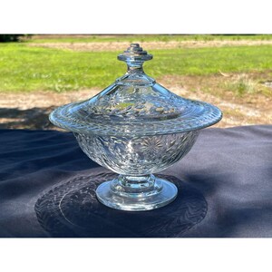 May include: A clear glass candy dish with a lid. The dish has a pedestal base and a round bowl with a lid. The lid and bowl are decorated with a starburst pattern. The dish is sitting on a dark blue surface.