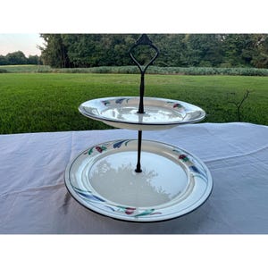 May include: A two-tiered serving stand with white plates featuring a floral design in blue, red, and green. The plates have a black trim and are connected by a black metal stand with a decorative handle.