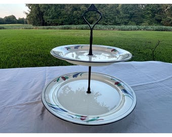 May include: A two-tiered serving stand with white plates featuring a floral design in blue, red, and green. The plates have a black trim and are connected by a black metal stand with a decorative handle.