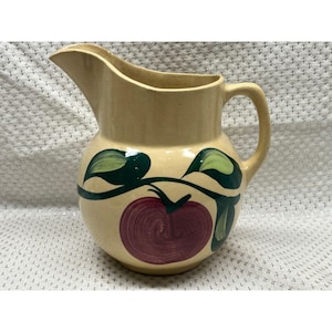 May include: A beige ceramic pitcher with a rounded body and a spout. The pitcher is decorated with a large, red tomato and green leaves. The handle is curved and the pitcher has a glossy finish.