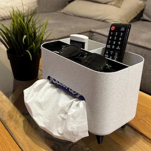 Pode incluir: Porta-lenços de papel branco com acabamento cinza salpicado. Tem um compartimento para um controle remoto, um telefone e óculos de sol. O porta-lenços tem um inserto para caixa de lenços de papel e um dispensador de lenços de papel retrátil.