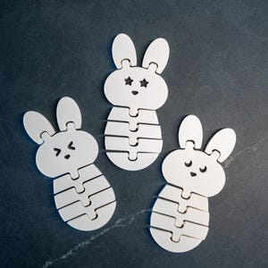 Könnte beinhalten: Drei weiße Hasenpuzzles mit ausdrucksstarken Gesichtern und segmentierten Körpern. Die Hasen haben unterschiedliche Gesichtsausdrücke, darunter sternförmige Augen, zwinkernde Augen und geschlossene Augen. Die Puzzles liegen auf einer dunkelgrauen Oberfläche.
