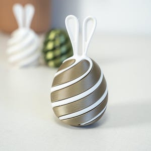 May include: A decorative Easter egg with a bunny ear design. The egg is gold with white stripes and white bunny ears. Other eggs are visible in the background, one white and one green.