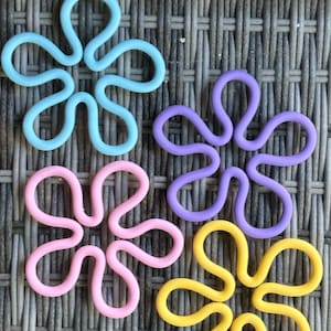 May include: Four colorful flower-shaped silicone pot holders in blue, purple, pink, and yellow. The pot holders are on a woven wicker surface.