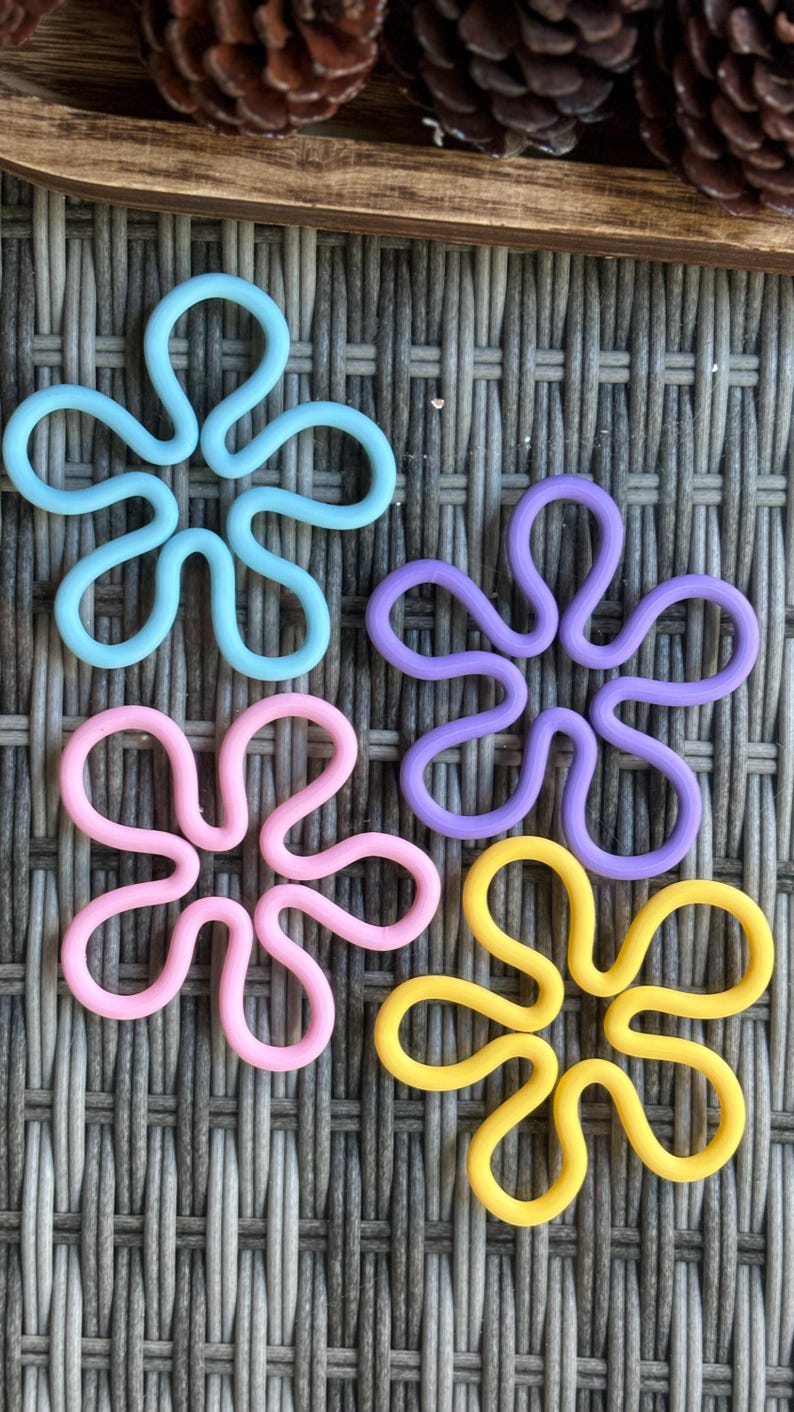 May include: Four colorful flower-shaped silicone pot holders in blue, purple, pink, and yellow. The pot holders are on a woven wicker surface.